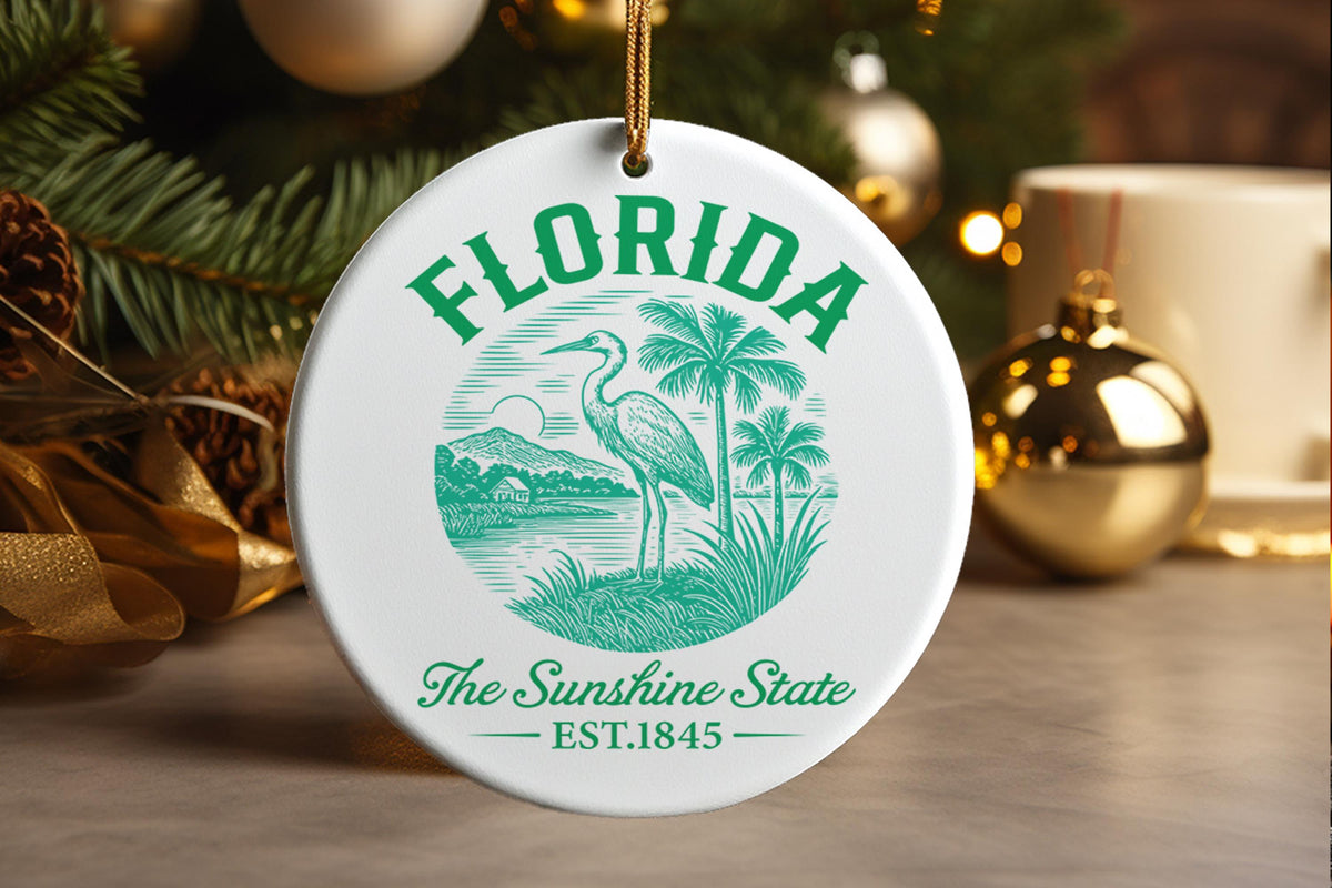 The design is a circular piece rendered in a vibrant monochromatic green tone against a white background, symbolizing the tropical seeing of Florida. Across the top, the word "FLORIDA" is prominently displayed. The central scene depicts a quintessential Florida landscape: lush palm trees frame a calm body of water where a tall wading bird stands guard. In the background, there are stylized silhouettes of distant landmasses or structures, evoking the state's varied environments. Beneath this scene, the inscr