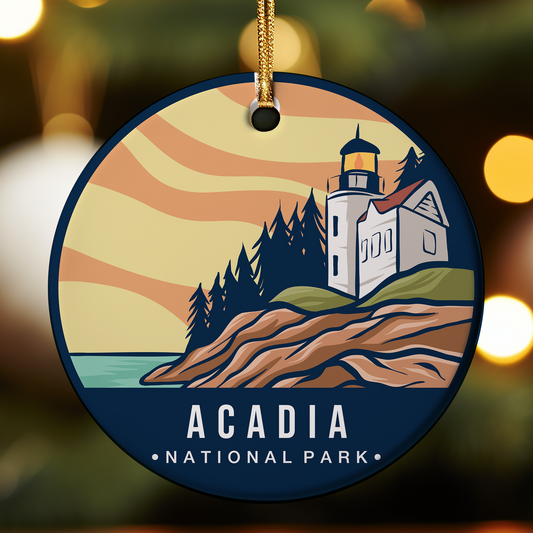 Round ceramic ornament featuring an illustrated scene of Acadia National Park in Maine, with a white lighthouse and keeper’s house on a rocky coastal cliff, surrounded by dark pine trees and overlooking the ocean under a warm, wavy sunset sky. The bottom of the ornament displays the text “ACADIA NATIONAL PARK,” and it hangs from a gold cord against a blurred festive background.