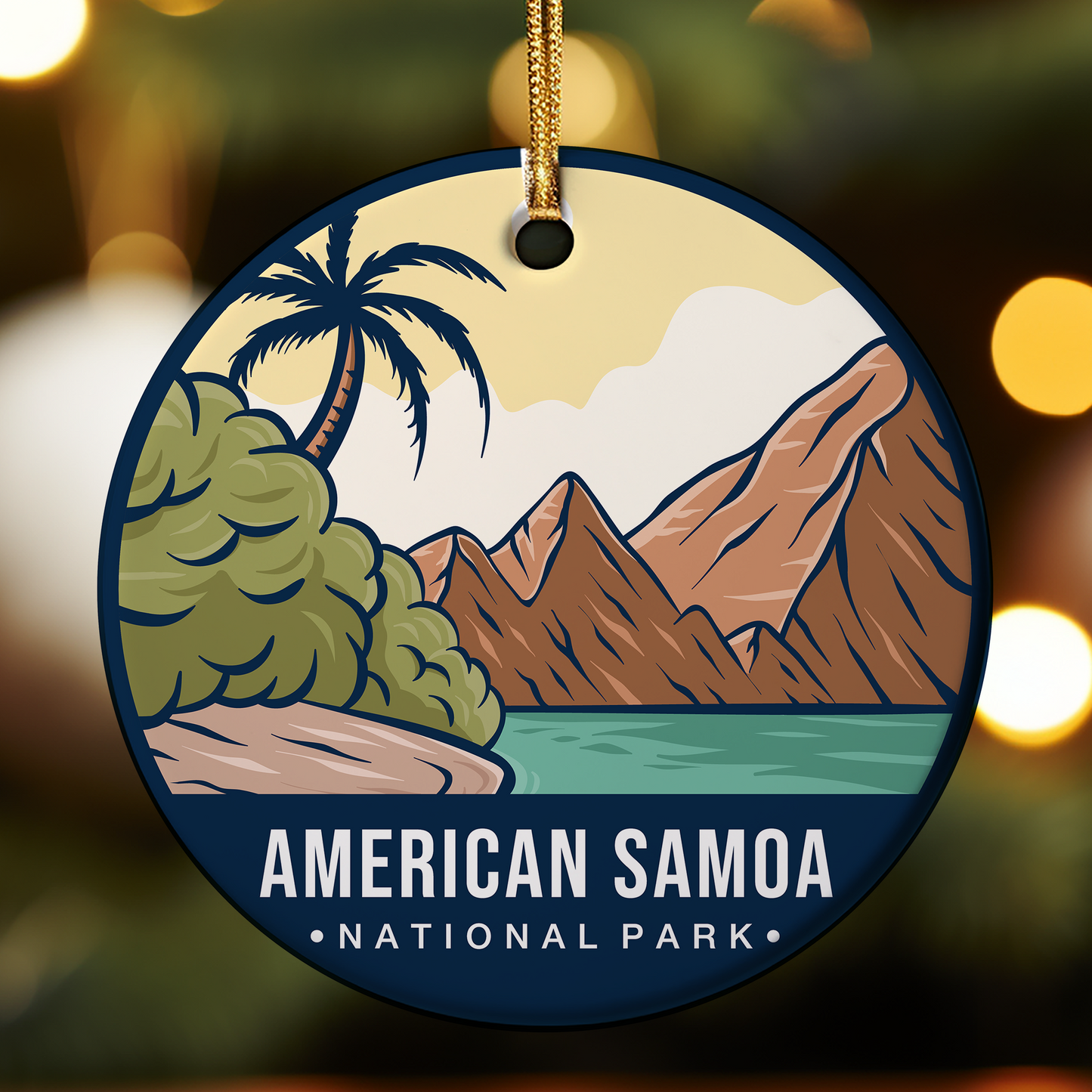 Round ceramic ornament featuring an illustrated tropical landscape from American Samoa National Park, with a palm tree and lush greenery beside a shoreline, turquoise water, and tall brown volcanic mountains under a soft yellow sky. The bottom of the ornament reads “AMERICAN SAMOA NATIONAL PARK,” and it hangs from a gold cord against a blurred festive background.