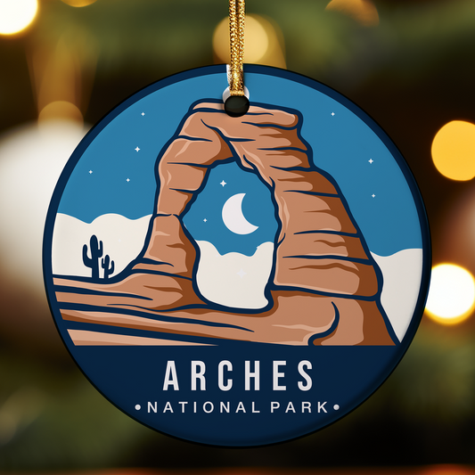 Round ceramic ornament featuring an illustrated scene of Arches National Park in Utah, with the iconic Delicate Arch rock formation in a desert landscape. A crescent moon and stars appear in a blue night sky above sandstone formations and a small cactus in the distance. The bottom of the ornament reads “ARCHES NATIONAL PARK,” and it hangs from a gold cord against a softly blurred festive background.
