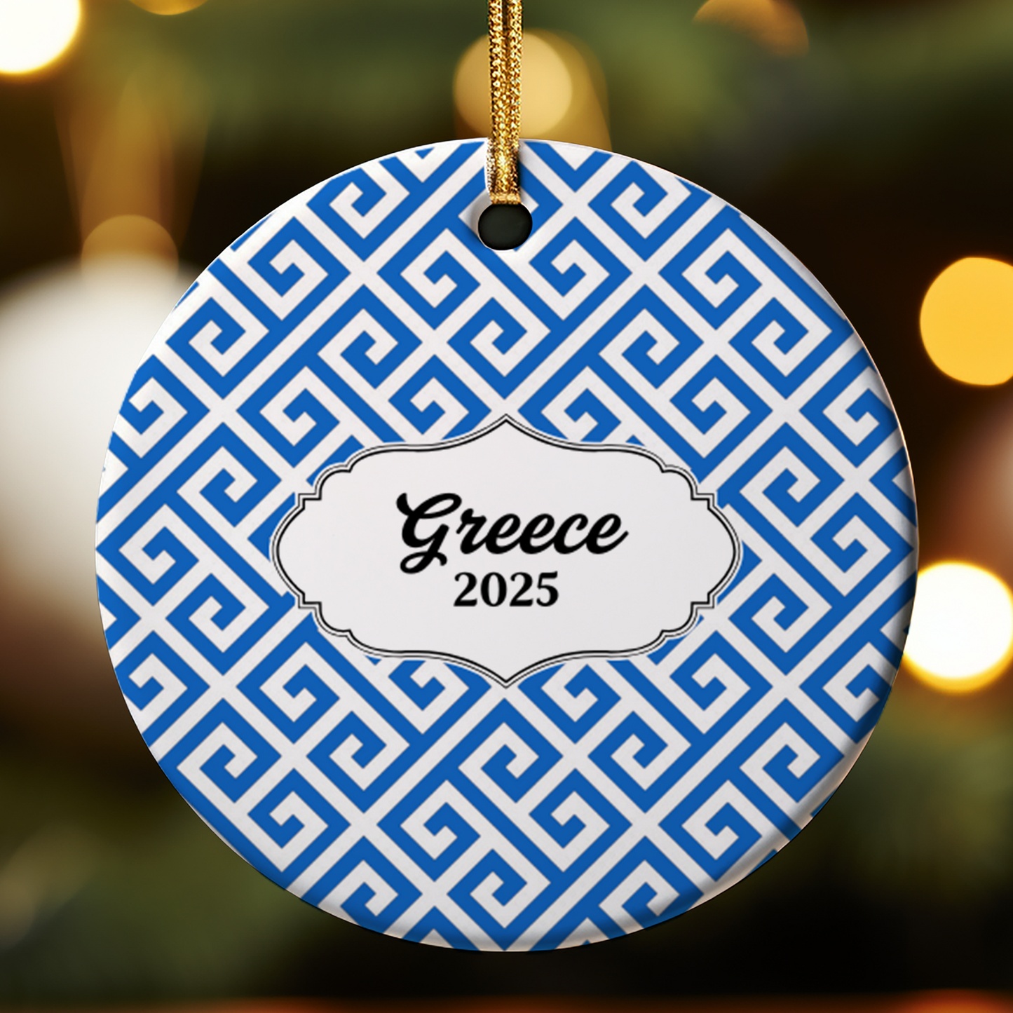 Decorative ornament with blue and white pattern and 'Greece 2025' text, hanging against a blurred festive background.