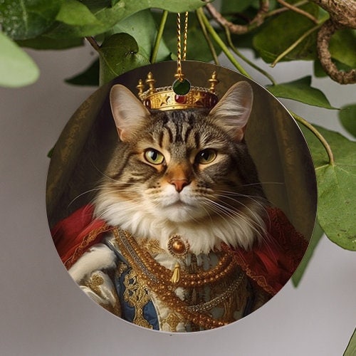 This holiday ornament displays a photorealistic image of a Mackerel Tabby cat wearing a golden crown studded with rubies in royal attire. Deep red velvet cloak and golden pearls adorn this adorable cat.