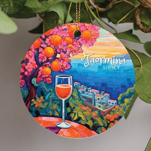 This holiday ornament shows a glass of wine on a table in the foreground during a warm afternoon sky. A steep cliff plunges toward the cove overlooking the turquoise waves of the Ionian Sea.