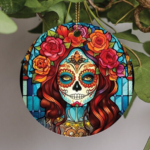 This holiday ornament is for The Day of the Dead Celebration. A stained glass design decorates the background. A long red haired sugar skull woman with orange floral eyes and big colorful flower in her hair sits in front.