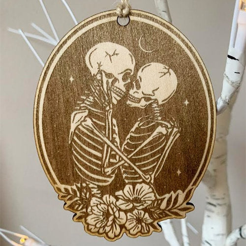 This holiday ornament shows two skeleton crossed lovers under a crescent moon starry sky, in an oval frame and flowers toward the bottom.