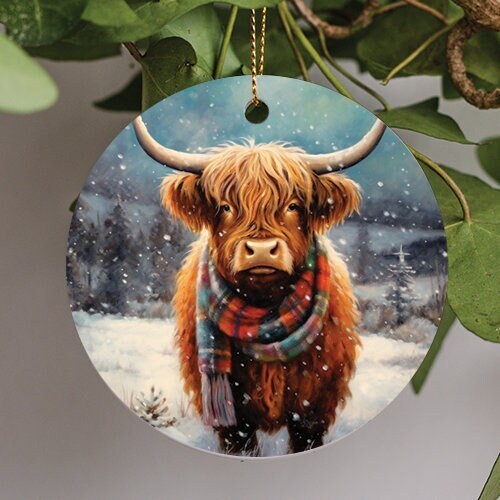 This holiday ornament shows an adorable long haired Highland Cow wearing a holiday inspired warm scarf on a snowy winter day.