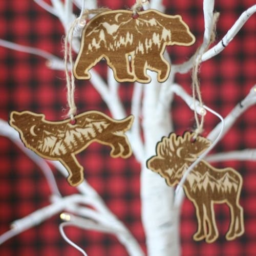 set of three ornaments of a moose, bear, and wolf made of wood. Silhouette of mountains are engraved into wood.