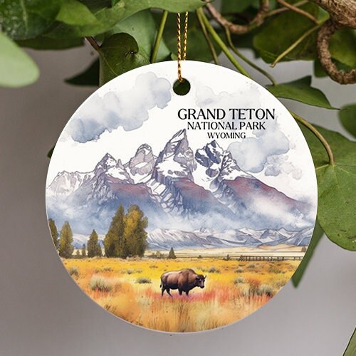 This ornament of Grand Teton National Park in Wyoming has a cloudy snow gray sky in a watercolor paint style. Mountains  snow capped tops. A lone bull stands amongst the long warm looking yellow and orange long grassy field.