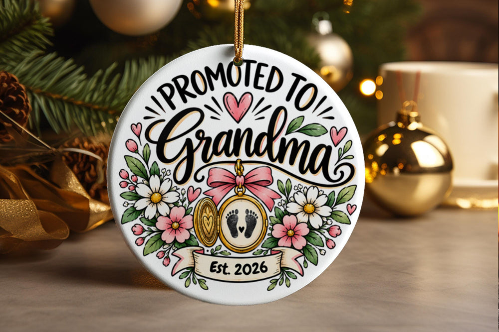A round white ceramic Christmas ornament hanging on a tree branch, featuring the text 'PROMOTED TO Grandma' with pink hearts, floral illustrations, baby footprints in a gold locket, and 'Est. 2026' on a banner.