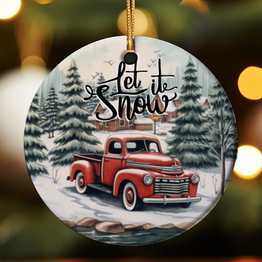 Round ceramic Christmas ornament featuring a vintage red farm truck in a snowy winter wonderland scene with snow-covered pine trees and a farmhouse in the background, with "Let It Snow" script text, hanging from a gold ribbon.