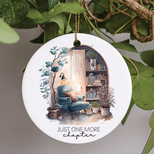 This Everyday Ornament is a watercolor illustration of a cloth wingback chair in a reading room. A window sits behind with sheer curtains, a wooden bookshelf sits off to the right with four shelves stuffed with books. Potted plants scatter the floor and a warm night light sits just above the wingback chair. A very peaceful serene setting with the text - Just One More Chapter underneath.